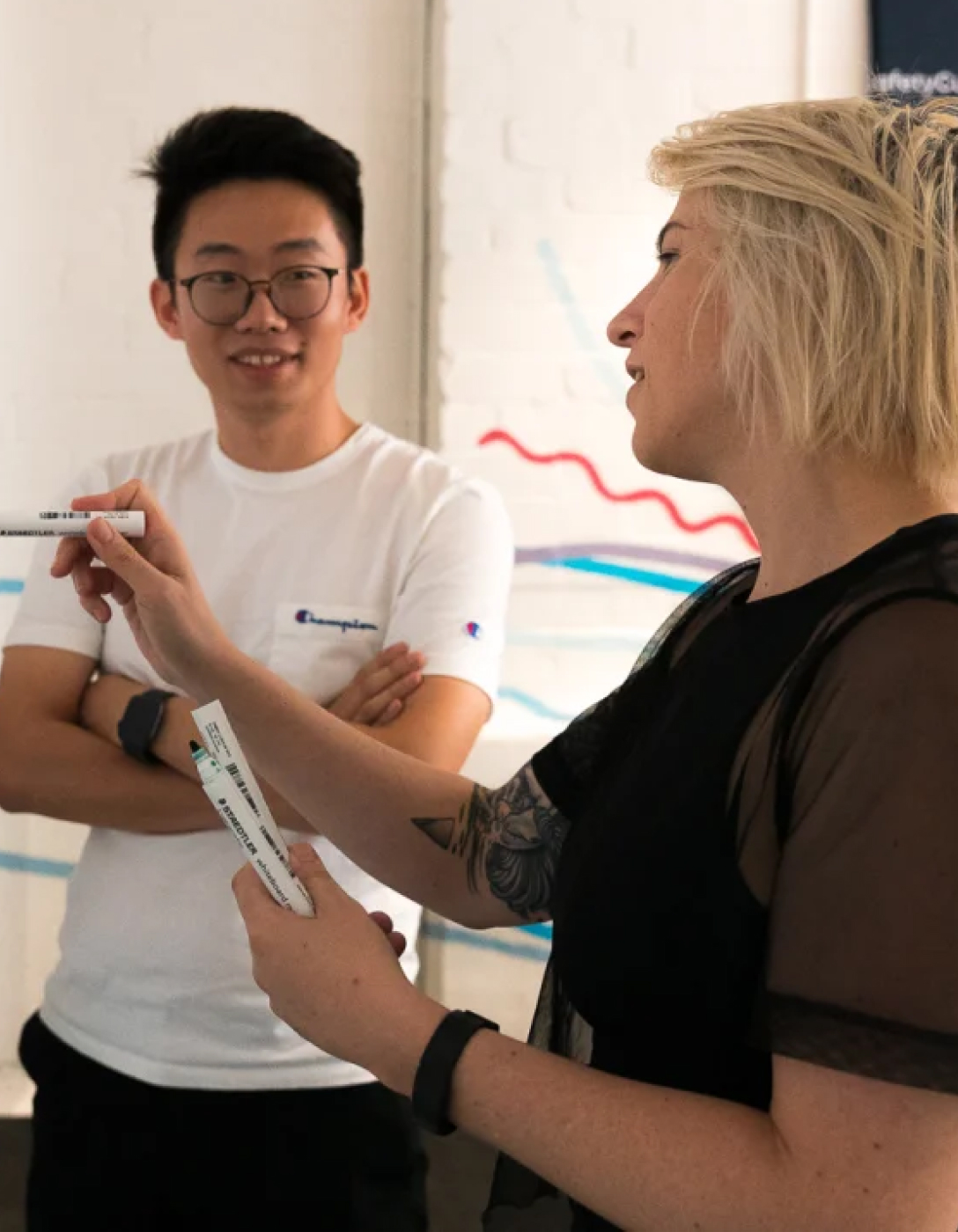 Two people in conversation, one in a white Champion shirt and one with blonde hair in black, both holding markers.